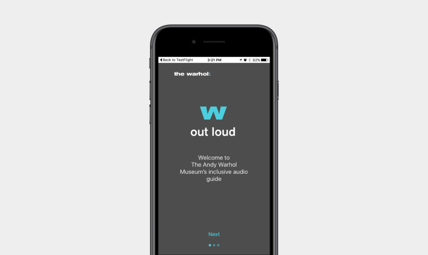A black iPhone centered on a light gray background. On the phone screen is a dark gray app welcome page branded "the warhol:" at the top. In the center, a large turquoise "w" appears above the title "out loud" in white text. Below it, the screen reads: "Welcome to The Andy Warhol Museum's inclusive audio guide." Near the bottom is a turquoise "Next" button with three small pagination dots underneath, indicating an onboarding sequence. The iOS status bar is visible at the top, including "Back to TestFlight," the time, and battery status.