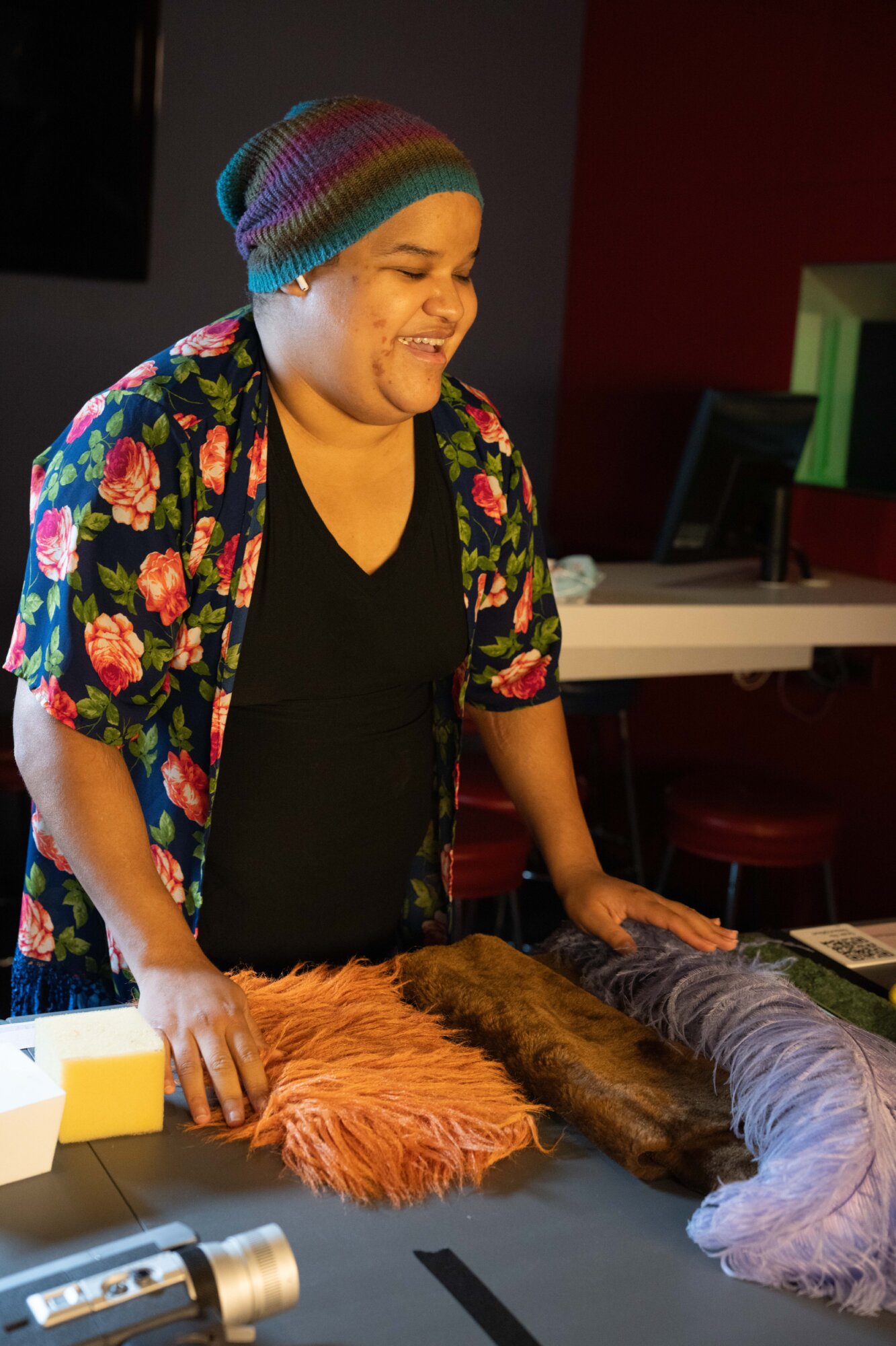 Nefertiti Matos Olivares, a Latina woman with medium skin tone, leaning over a table and smiling wide with her mouth open, her eyes closed, as he touches long orange fur and a purple ostrich feather. The table also has short brown fur, foam blocks and a tactile QR code near the edge. Nefertiti is wearing a colorful striped knit beanie, a dark shirt, and a blue floral outer shirt with bright pink and orange flowers. A white earbud peeks out of one ear. The background is a dark exhibition space, with dark walls, red seating, and warm lighting.