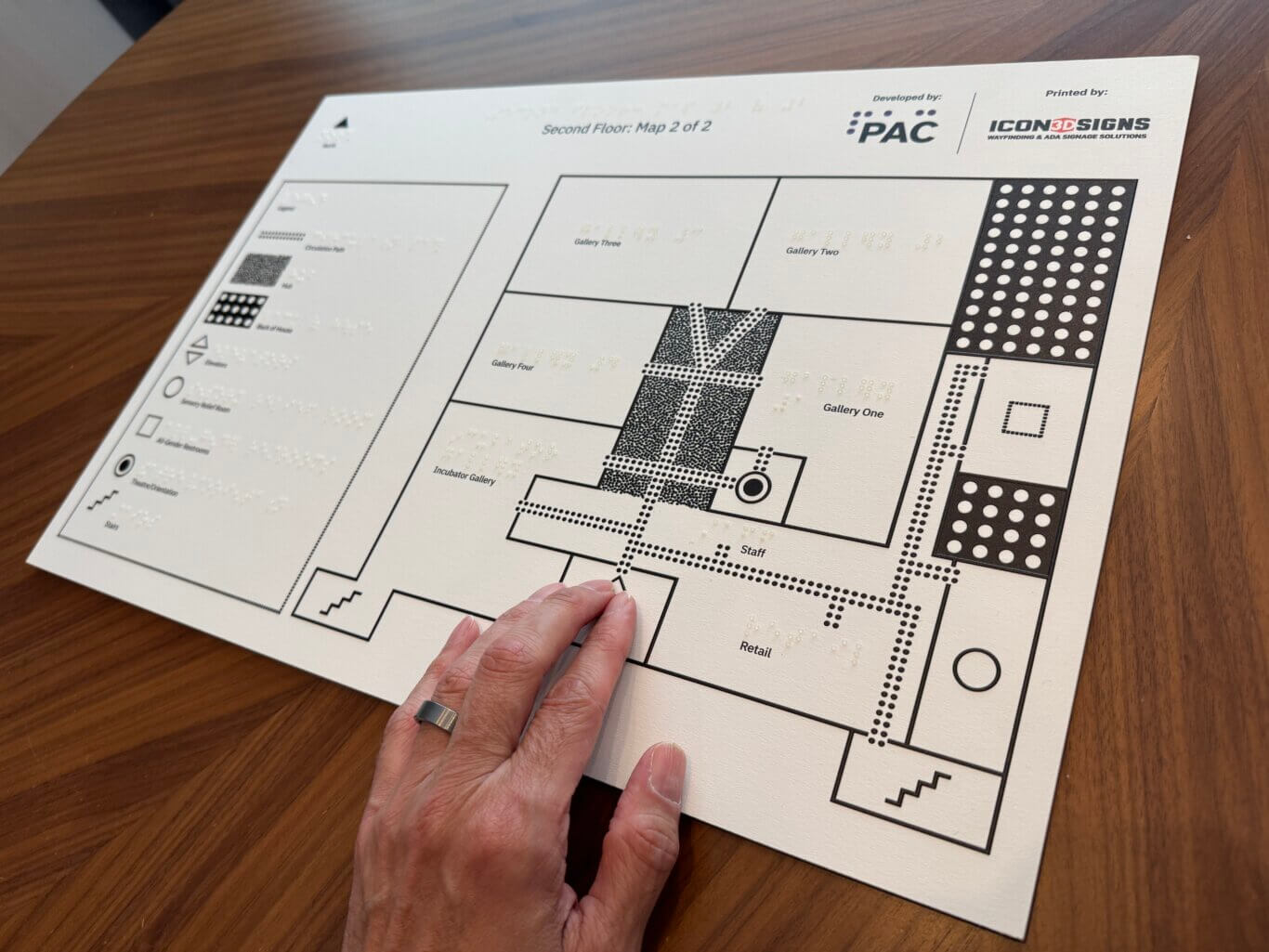 A close-up of a large printed tactile wayfinding map titled "Second Floor: Map 2 of 2." The map includes raised Braille text, bold black room outlines, dotted tactile pathways, and labeled spaces such as Gallery One, Gallery Two, Gallery Three, Gallery Four, Retail, Staff, and Incubator Gallery. A legend appears on the left with symbols for exits, restrooms, stairs, and other features. At the top are logos for PAC and ICON3DSIGNS. A light skin toned hand wearing a wide metal ring touches the lower part of the map. The map is placed on a warm wooden tabletop.