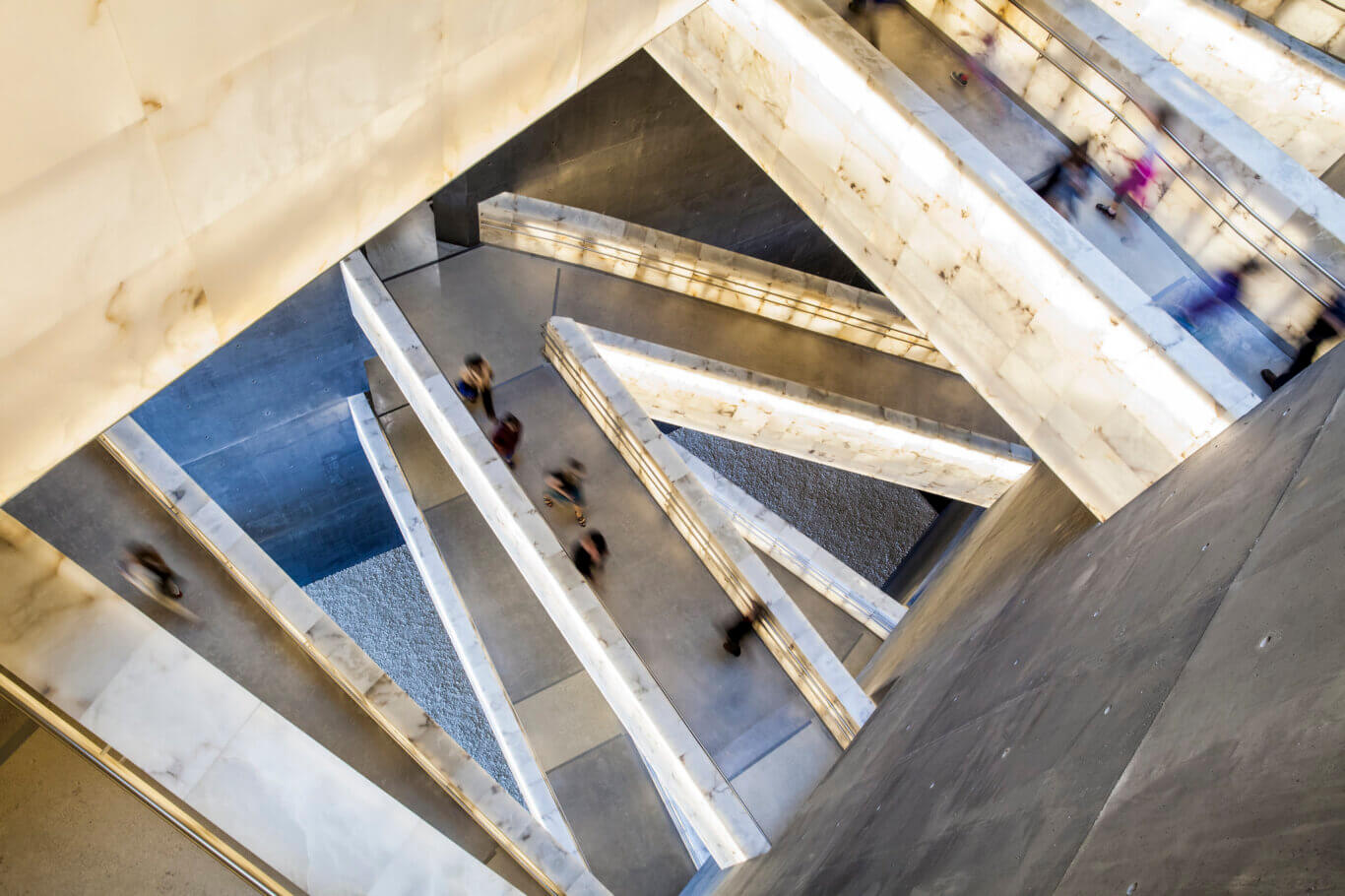 An architectural interior photographed from above, looking down through several intersecting levels of ramps or stairways. Large illuminated white stone or marble panels create bright geometric lines, contrasting with gray concrete walls and floors. Metal railings run along the walkways, and several people appear as motion-blurred figures, suggesting movement through the space. The composition is highly angular and abstract.