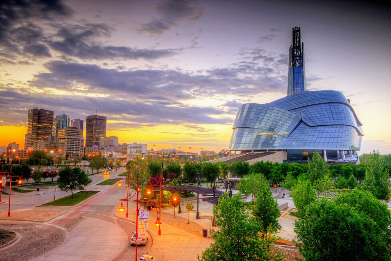 A panoramic urban scene at dusk, with a dramatic sky filled with purple-gray clouds and warm golden light near the horizon. On the right is a striking modern glass building, the Canadian Musuem for Human Rights, with curved, reflective panels, that resemble a shell or layered seed pod, and a tall tower rising from its roof. On our left, downtown high-rises are lit by the fading sunlight. In the foreground, tree-lined roads, sidewalks, red streetlamps, and light trails from passing cars.