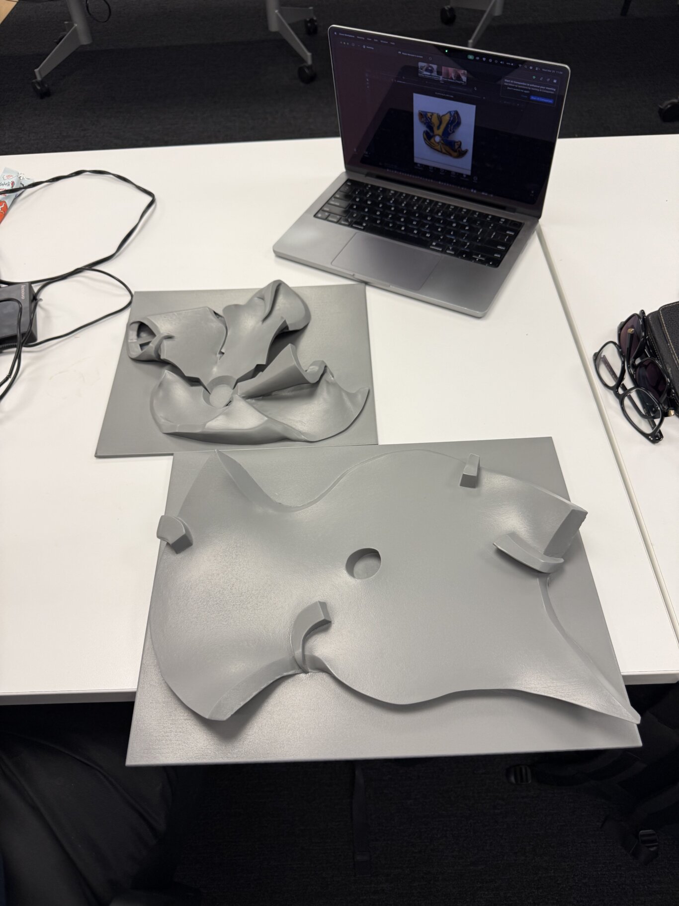 A meeting room table with two gray tactile objects laid out on flat base panels and a laptop behind them. The pieces have curved, abstract shapes with raised edges, cutouts, and smooth sculpted surfaces. An open laptop sits toward the back right, displaying a dark interface with an image of a similar object in full color on the screen. Around the table are loose cables, a small device or adapter, eyeglasses, and a partially visible snack wrapper.