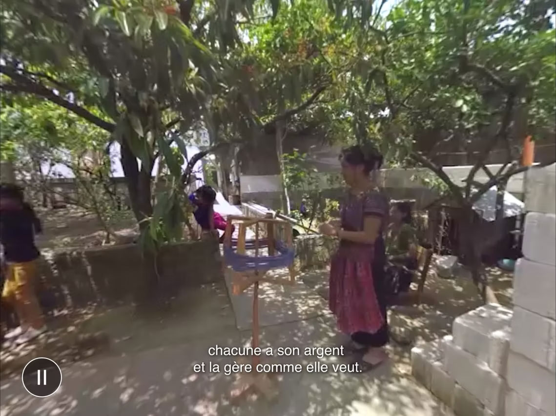 A still from a video of a woman with medium brown skin tone and a purple and pink dress in a sunny courtyard or garden area shaded by dense trees and branches. The woman is slightly blurred but can be seen interacting with an upright wooden structure where she may be spinning yarn around it. White blocks or bricks are stacked on the right side, and sunlight filters through the leaves, creating dappled shadows on the ground. A French subtitle at the bottom reads: "chacune a son argent et la gère comme elle veut." There is also a pause icon in the lower-left corner, suggesting this is a paused video.