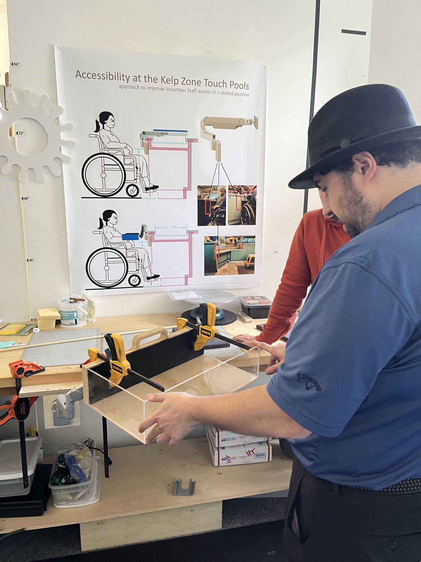 Taken in a design or fabrication workspace, Sina Bahram, a Persian man, holds a prototype for an accessible kelp zone touch pool. On the wall is a large poster labeled "Accessibility at the Kelp Zone Touch Pools," with diagrams showing a wheelchair user positioned next to a touch-pool counter and proposed design modifications for seated access. The poster includes measurement references and small photos of an existing exhibit area. In the foreground, Sina holds a transparent acrylic box or tank-like prototype. Several yellow and orange clamps hold parts in place, and there are tools, cutting mats, wood pieces, containers, and supplies scattered around the table.