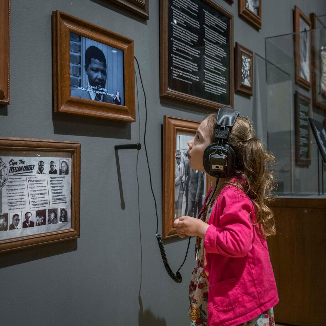 A young girl with pale skin in a bright pink jacket standing close to a museum wall, wearing oversized black headphones connected by a cord to an audio guide. She is looking up at a small framed video screen displaying black-and-white footage of Nelson Mandela with French subtitles. Around the screen are several wooden-framed exhibits, including historical photographs, text panels, and documents related to freedom, civil rights, and anti-apartheid history.