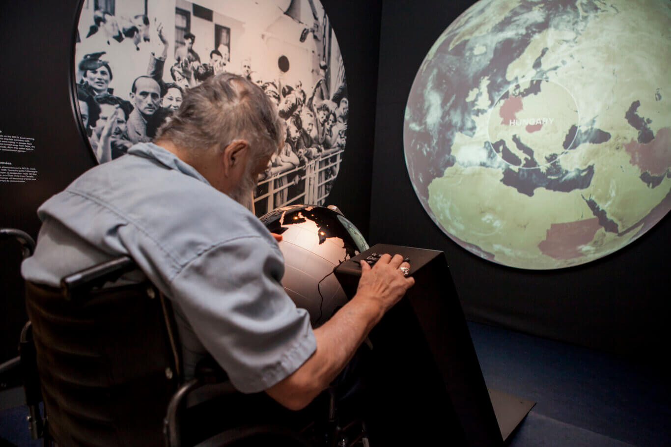 An older man with light skin tone seated in a wheelchair, leaning forward to use a universal keypad (UKP) next to a globe at an interactive museum exhibit. In front of him is a small globe-like display, while a large circular projection on the wall shows a map of Europe with "HUNGARY" highlighted. To the left, a large black-and-white historical photograph of a crowd is displayed.