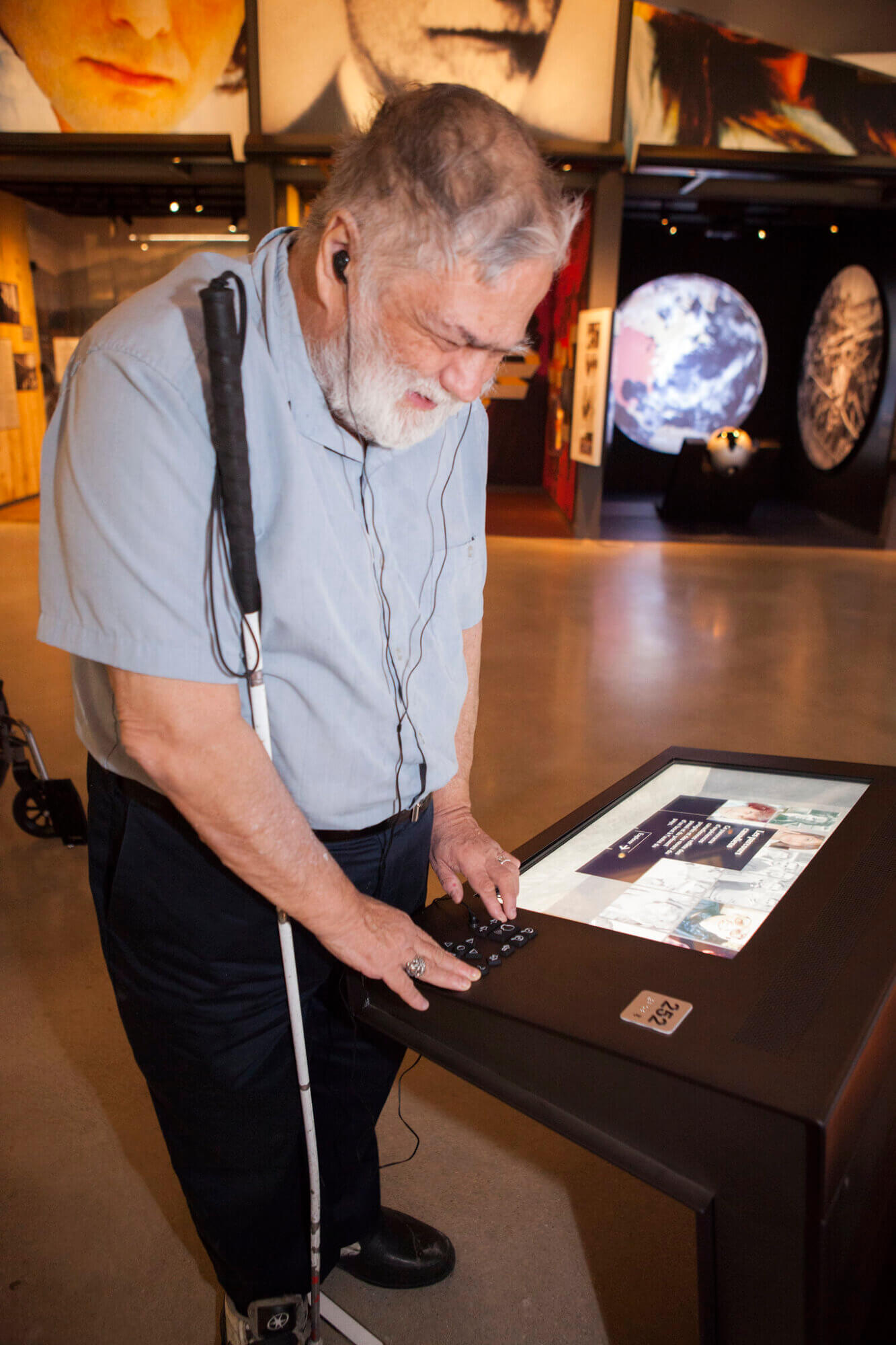 An older bearded man with light skin tone standing at a digital museum display, leaning over an interactive touchscreen kiosk and pressing buttons on a universal keypad (UKP). He is wearing earphones plugged into the kiosk and holding a long white cane. Above the keypad is a small silver tactile square with the number 252 in raised print and Brailled. The museum space around him is dimly lit, with large portrait panels and space-themed displays visible in the background, including an image of Earth and a spacecraft model.