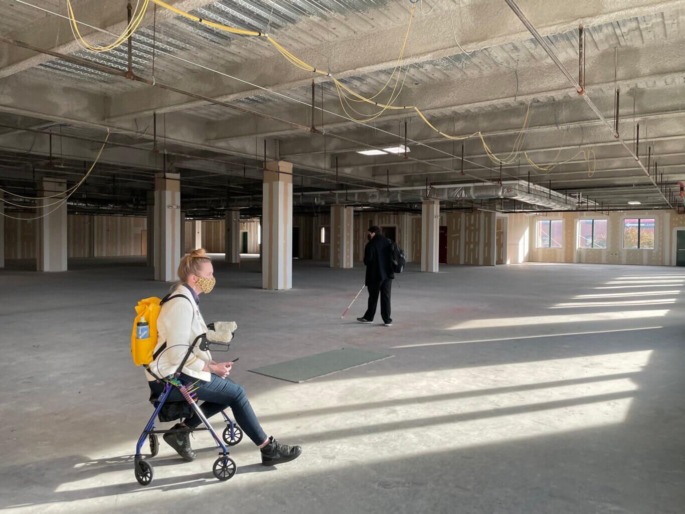 Two people in a large, open indoor space under construction. The ceiling is unfinished, with exposed pipes, sprinkler lines, ductwork, hanging electrical cables, and yellow wiring loops. The walls are mostly bare drywall with visible joint compound, and there are many square columns throughout the space. Sunlight streams in from windows on the right, casting long striped shadows across the concrete floor. In the foreground, Robin Marquis, a white nonbinary person wearing a leapord print maks sits in profile on a blue rollator, wearing a yellow backpack and holding a phone. In the background, Sina Bahram, a Persian man, wearing a black mask and dressed in dark clothing walks towards the back area with a long white cane.