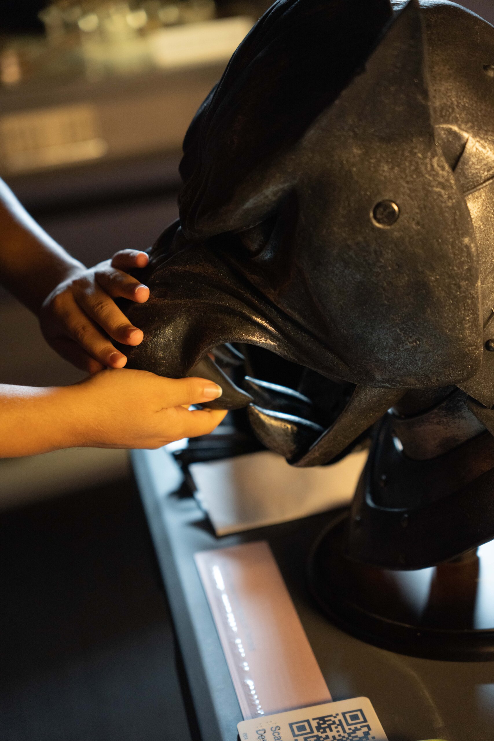 A close-up of small hands with medium skin tone touching the snout of a black helmet in the shape of a stylized hound or dog with bared teeth. The hands touch the open mouth with four massive sharp teeth, and the wrinkled nose as if the armored mask is in mid snarl. The rest of the mask is made of angular plates that extend down the neck. A blurry label with print and Braille and a tactile QR code is partially visible near the edge of the table.