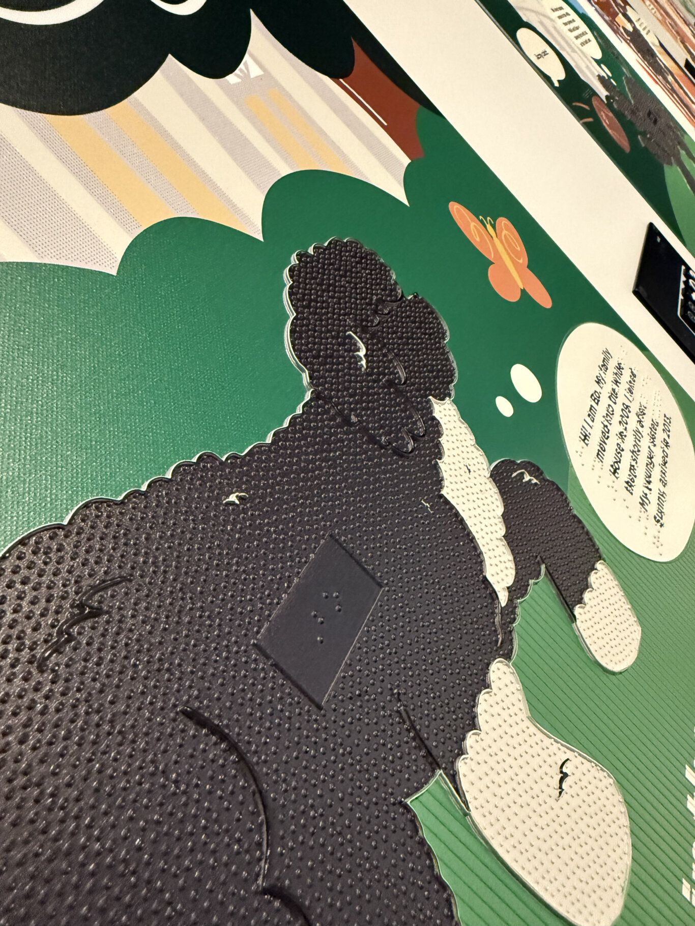 A tight, angled close-up of a glossy, textured illustrated surface of a tactile dog in a children's story book. The tactile image depicts a black and white dog with raised lines and varied texture mapped to the different colors. In the middle of the dog is a Braille label. On the right side, there is a small orange butterfly graphic on the green background, and a white speech-bubble-like area with partially visible black text and Braille.