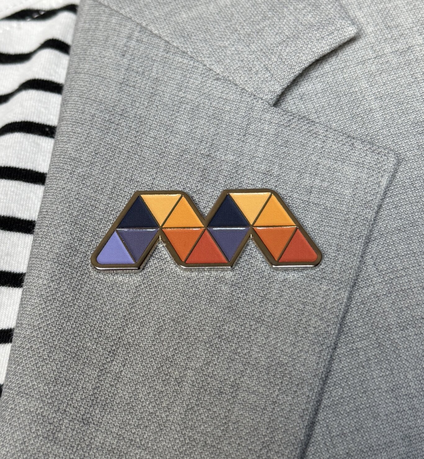 A close-up of a geometric enamel Mosaic “M” pin attached to a light grey, textured blazer or jacket. The pin is made of triangular sections in orange, yellow, red, navy, purple, and light blue with dark borders. Along the left edge, part of a black-and-white striped ribbed fabric is visible. The gray material has visible seams and a soft, slightly fuzzy texture.