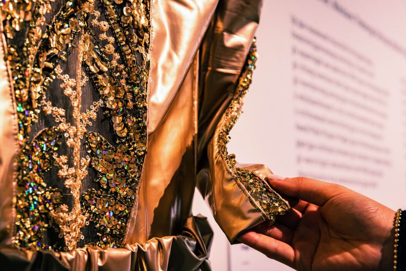 A tight, detailed close-up of someone with medium dark skin tone touching the gold lamé sleeve of a richly decorated gold outfit. Black mesh panels are covered with gold sequins, beads, floral embroidery, and reflective embellishments that catch the light in rainbow-like highlights. Shiny metallic gold fabric frames the embroidered sections and the background is softly blurred.