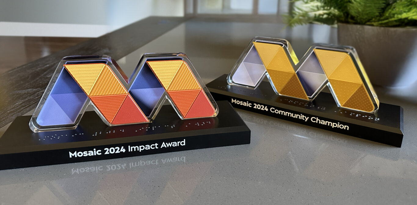 Two geometric Mosaic awards on a light-grey countertop, each shaped like a stylized “M” in blue, grey, gold, and orange panels. The triangles have a slightly textured surface that increase in density as the colors increase in saturation. They sit on black bases with engraved titles—“Mosaic 2024 Impact Award” and “Mosaic 2024 Community Champion”—and braille along the front.