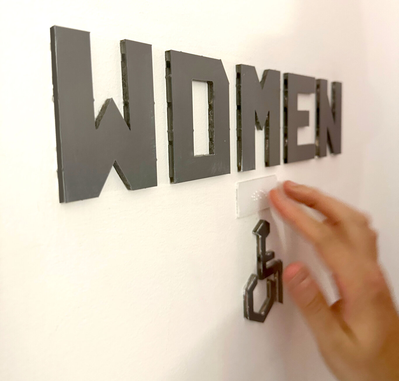 A close, angled view of thick gray block letters mounted on a white textured wall spelling the word, "WOMEN." Below the sign, a blurred hand with light skin tone touches Braille with the Blue Bob wheelchair access symbol below the Braille.