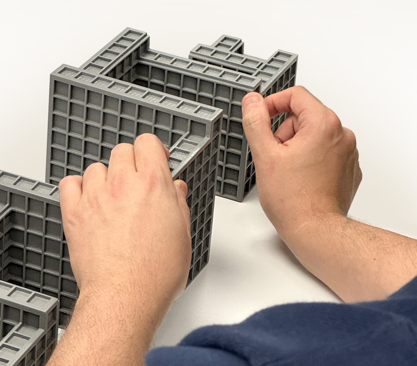 A close-up of a person with light skin tone using both hands to handle touch a tactile model of many cubes stacked in grid like paterns. The object has a repeating square grid pattern on its walls and top edges, resembling a miniature building or construction system. The structure appears hollow in the center, with multiple connected rectangular sections.