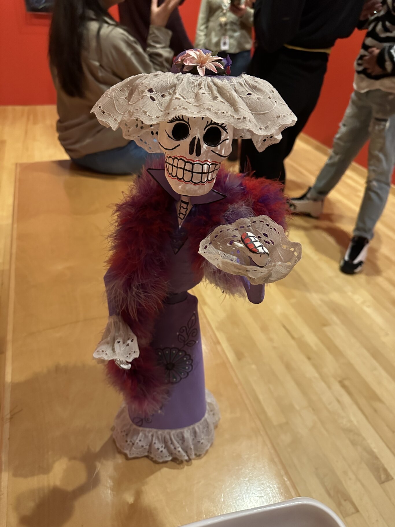 A paper maché sculpture of Catrina, a skeleton of a woman associated with Day of the Dead. She is about the size of an upright baby doll, has a painted skull face with large black eye sockets, a wide toothy grin, and decorative facial lines. She wears a large white lace hat topped with a small pink flower, a purple dress with black floral designs, and a fluffy red-purple feather boa. One of her arms is raised towards us with lace wrist cuffs, that match her hat and the edge of her dress. The background is softly blurred, showing people in a gallery-like room with a bright red wall and wooden floor.