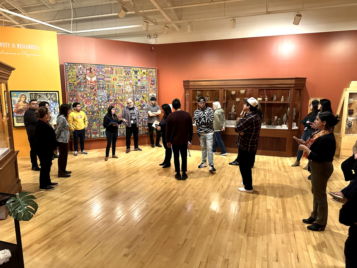 A large group of mostly Latino people with light to medium skin tones and various genders stand in a museum exhibition space with polished wooden floors and brightly redish brown and yellow walls. Behind them is a large, vibrant grid-like artwork filled with colorful symbols and patterns, while nearby glass cases display sculptures, masks, and other artifacts. A yellow wall on the left includes partial text referencing Mesoamerica.