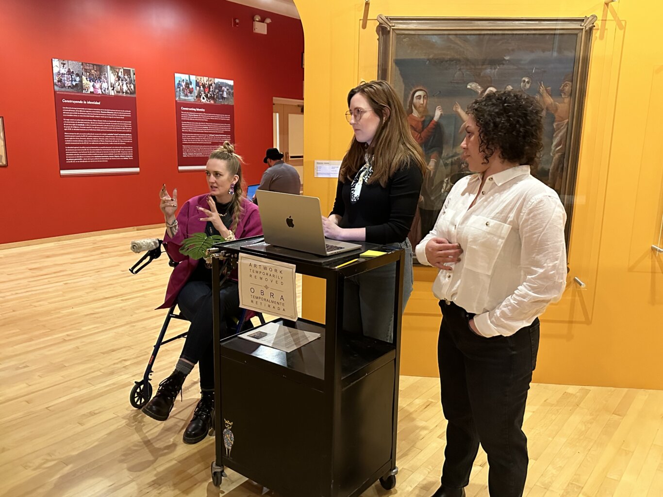 Several people are gathered near a black rolling cart holding an open Apple laptop and papers inside a colorful gallery, as if presenting to people out of view. On our left, Robin Marquis, a nonbinary white femme, gestures with their manicured nails in mid sentence sittin on a rollator. In the middle behind the rolling cart and laptop stands Maria Braswell, a white woman, and on our far right, Sara Mikail, a white woman, stands watching Robin. In the background, large framed paintings hang on the bright red and yellow walls, and informational exhibition panels titled "Construyendo la identidad" and "Constructing Identity" are visible.