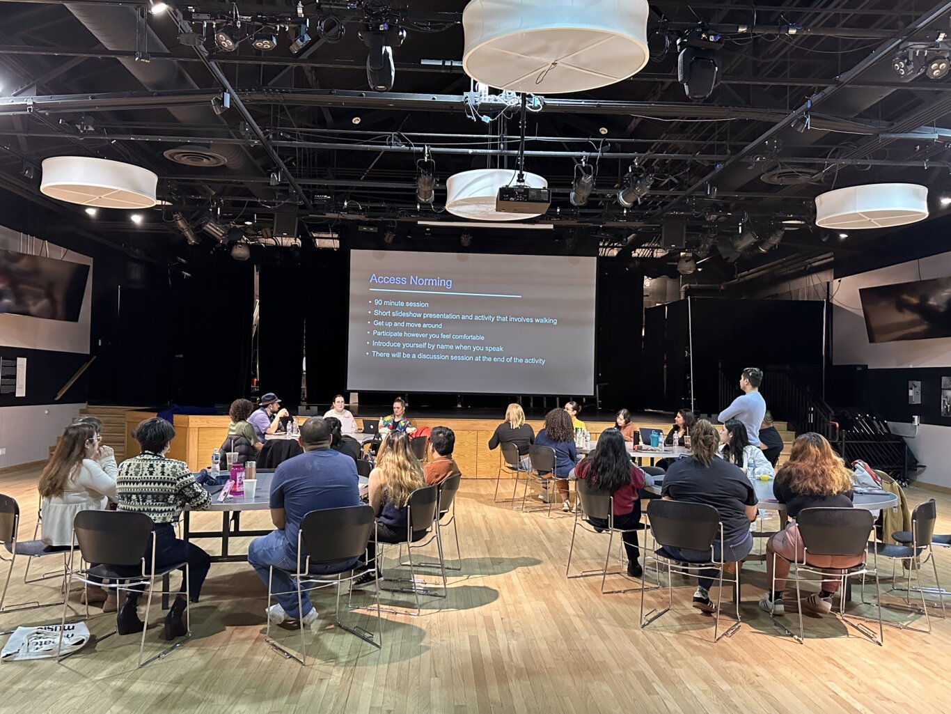 An indoor workshop in a spacious auditorium-like room with a wooden floor. Attendees are seated on black chairs around round tables, facing a stage area and a large projection screen. The visible slide is titled "Access Norming" and includes the bullet points, "90 minute session," "Short slideshow presentation and activity that involves walking," "Get up and move around," "Participate however you feel comfortable," "Introduce yourself by name when you speak," and "There will be a discussion session at the end of the activity." Several people are sitting near the front as facilitators, from our left to right, Sina Bahram, a Persian man, Maria Braswell, a white woman, and Robin Marquis, a nonbinary white femme. Overhead, the ceiling is filled with exposed beams, stage lights, cables, projectors, and large white circular hanging light fixturesl.