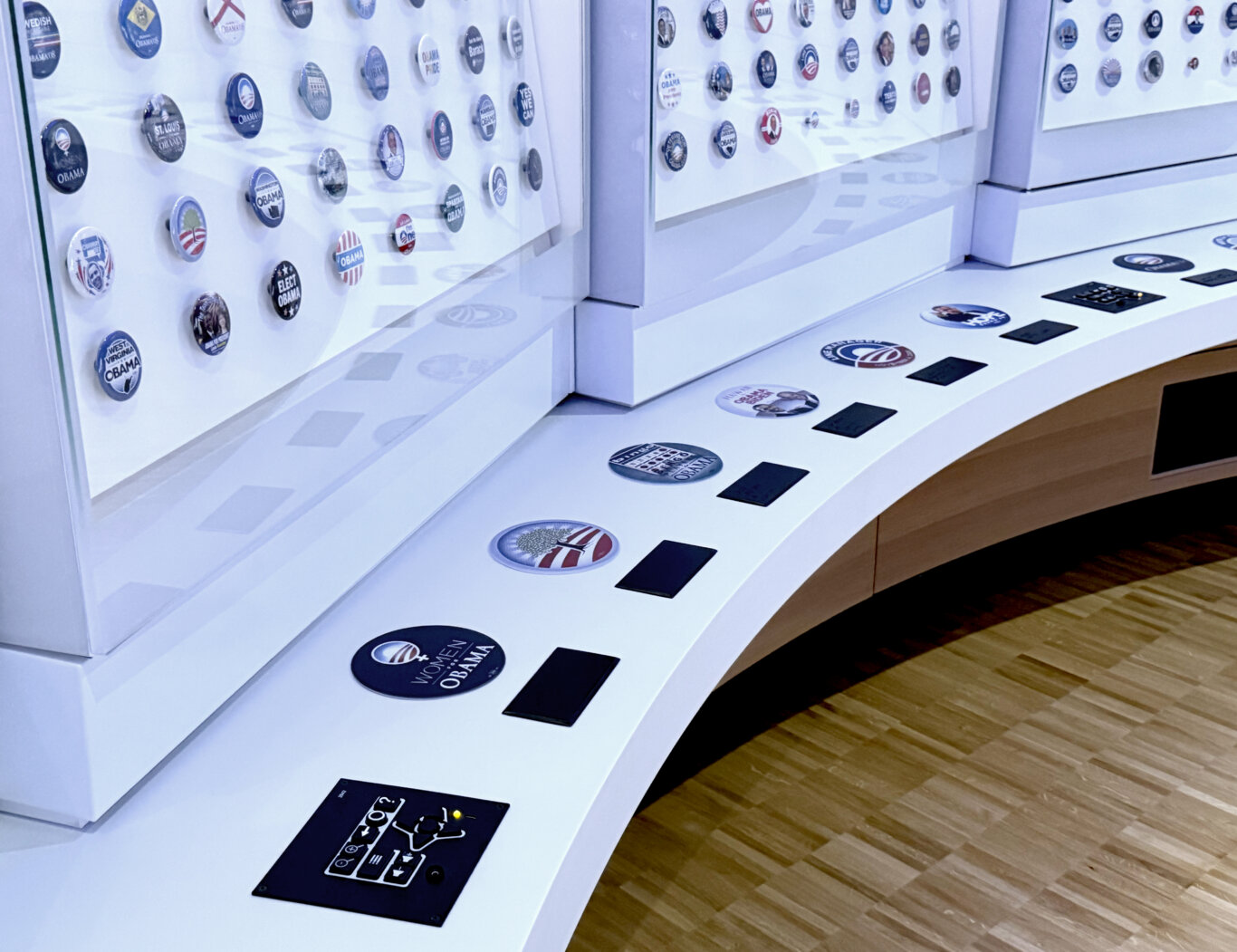 A bright, white exhibit display with rows of circular campaign buttons mounted behind glass and on slanted display surfaces.Underneath the cases on a ledge are large tactile versions of select buttons with black Brialle labels underneath. At the begining of the display, on our left is the UXP, a tactile keypad. Many of the pins feature Barack Obama campaign designs, slogans like "Yes We Can," and state or group-specific Obama memorabilia.
