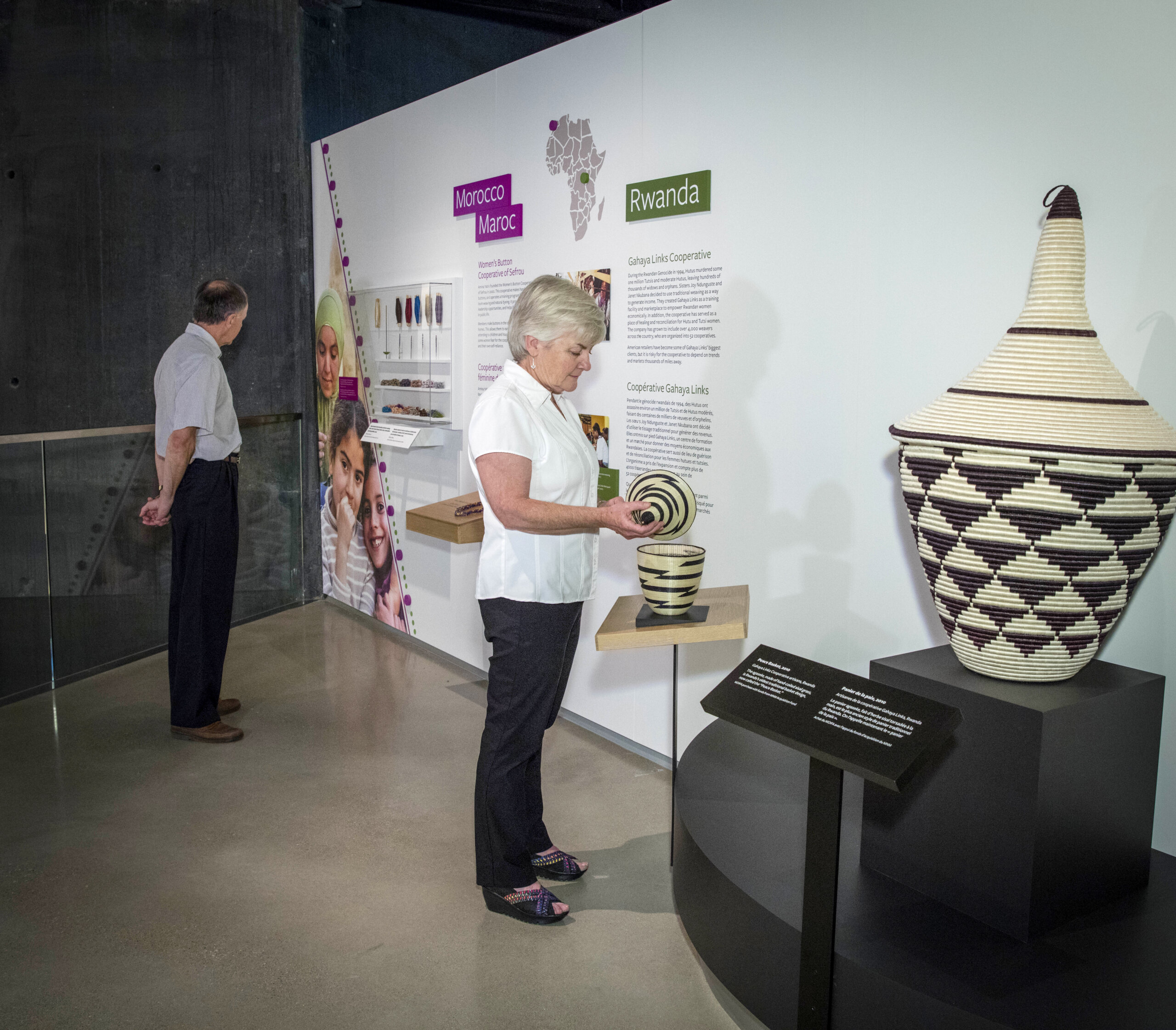 A museum exhibit with informational panels labeled "Morocco / Maroc" and "Rwanda." A map of Africa highlights locations, and wall text describes the Gahaya Links Cooperative in Rwanda with large woven baskets with black and cream patterns displayed on pedestals on our right. A woman with light skin tonepicks up the lid of a small basket that looks like a smaller version of the towering basket on our left. The Morroco exhibit features a large photo of smiling children with light brown skin tone arm in arm and a base with jewlery. The space has a modern, minimal design with white exhibit walls, dark structural elements, and polished concrete flooring.