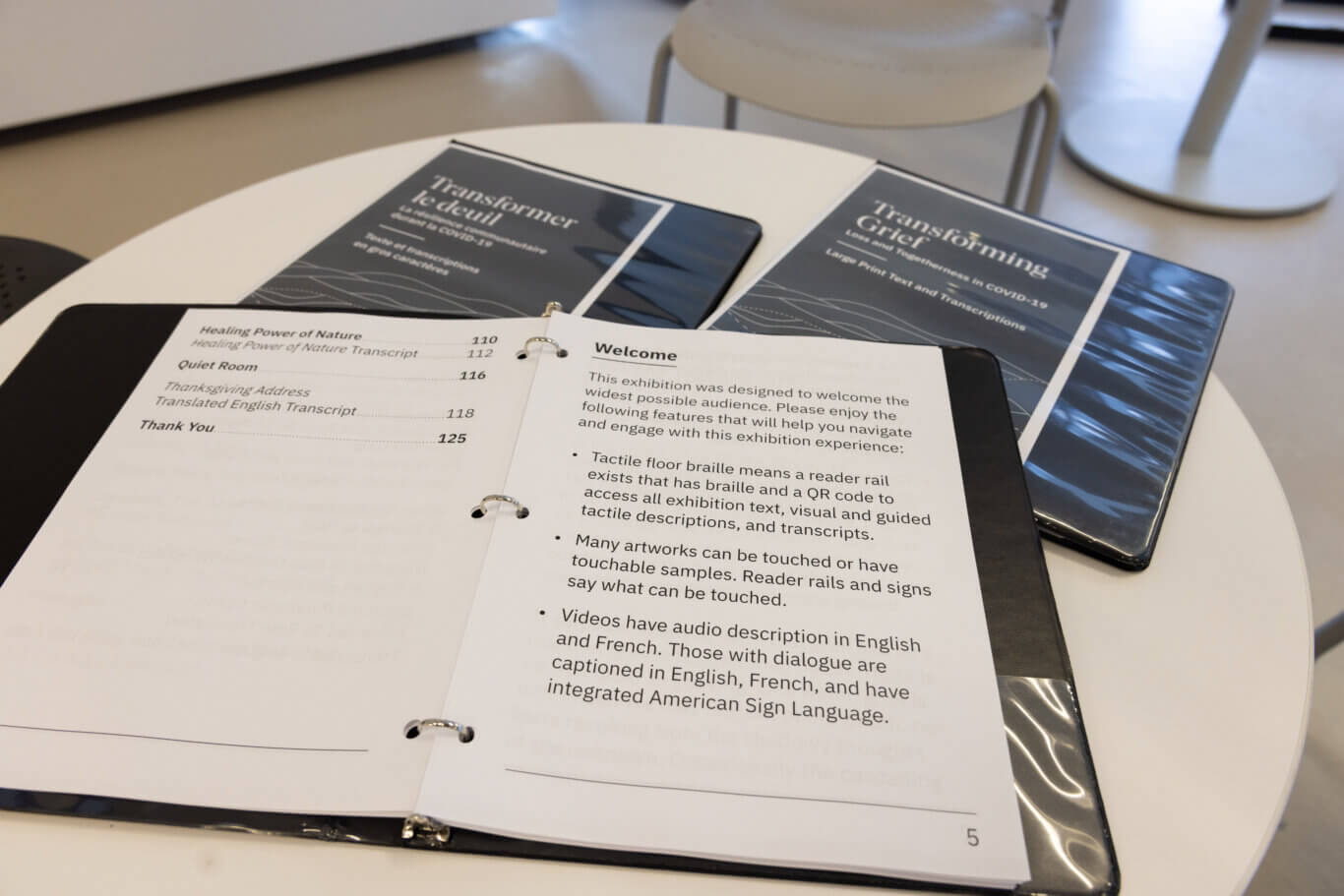 A close-up of printed exhibition materials in large print spread across a white round table. In the foreground, an open ring binder displays a "Welcome" page with large text describing accessibility features, including tactile floor braille, QR codes, touchable samples, audio description, captions, and American Sign Language. The opposite page appears to be a table of contents listing sections such as "Healing Power of Nature," "Quiet Room," "Thanksgiving Address," and "Thank You." Behind the binder are several dark-covered booklets titled "Transforming Grief," with subtitles referencing COVID-19, grief, mourning, and transcripts. The background is softly blurred, showing chairs and tables in a bright indoor setting.