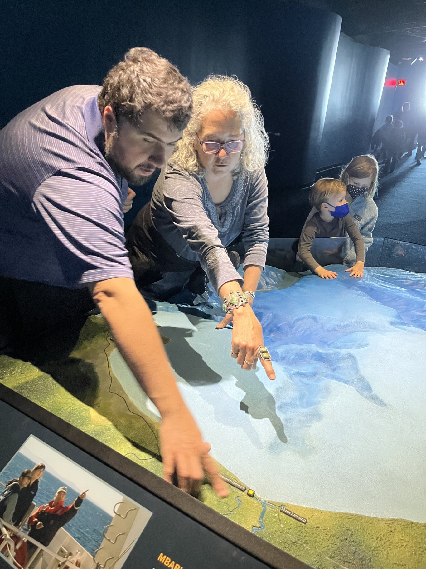 Two people lean over an aquarium-style exhibit with a large illuminated relief map featuring water, coastline, and labels such as "Research Institute" and "Moss Landing." On our left, Sina Bahram, a Persian man, and Beth Redmond-Jones, a white woman, reach across the display pointing and looking closely at something on the map. In the background, two small children with light skin tone wearing face masks are also touching the relief map.
