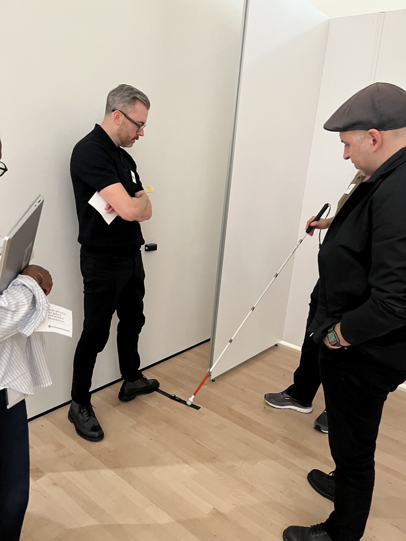 A few people stand in a small indoor room watching someone touch a tactile floor strip with their long white cane. On our left, Rob Itri Vincent, a Metí man is wearing all black with his arms crossed, holding a sheet of paper and looking downward. His foot is resting on a strip of black tape on the floor that is being touched with the tip of a long white cane, held by someone on our right, who's blocked by Corey Timpson, a white man wearing all black. Corey is also looking at the floor strip.