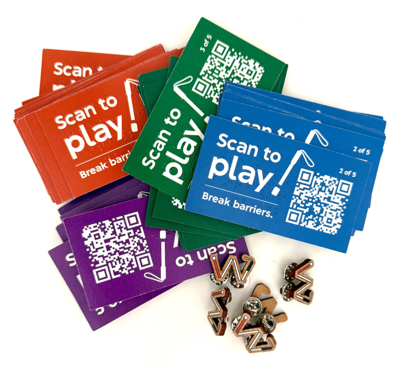 Overlapping piles of glossy rectangular cards in red, green, blue, and purple. The cards display clear Braille and large white text reading "Scan to play! Break barriers," along with a prominent QR code and small numbering such as "2 of 5" and "3 of 5." The cards are spread diagonally across a white background. In the lower-right area, small wooden badge-like pins of a long white cane partially unfolded into the shape of an "M" are scattered next to the cards.