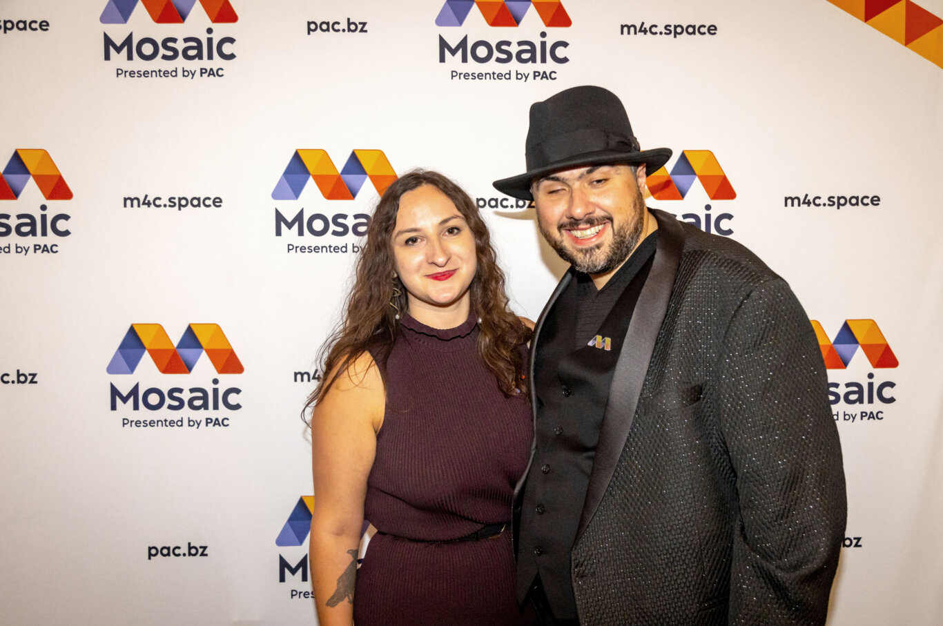 Two people pose in front of a white step-and-repeat backdrop for "Mosaic," featuring repeated colorful geometric M-shaped logos in orange, yellow, red, blue, and purple. Ruth Starr, a white woman, and Sina Bahram, a Persian man, smile at us wearing formal attire. On our right, Sina is wearing a black fedora and sparkly black suit jacket. Ruth is wearing a sleeveless burgundy dress and red lipstick. The backdrop includes readable text such as "Mosaic," "Presented by PAC," "m4c.space," and "pac.bz."