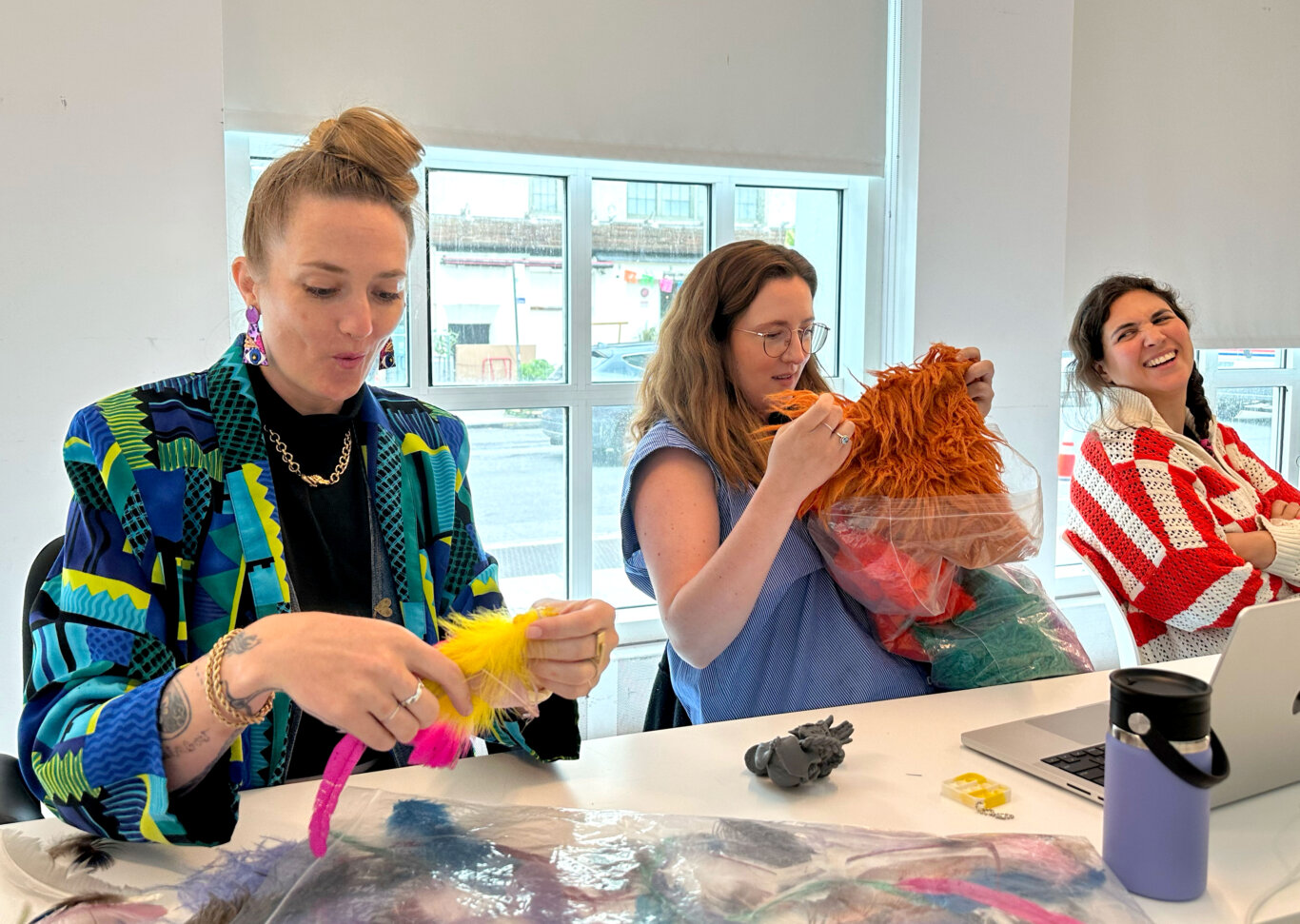 Three people sit at a table enthralled in touching muppet materials. Starting on our left, Robin Marquis, a white nonbinary femme holds a yellow and pink feather with their mouth pursed in mid squeal, in the middle, Maria Braswell, a white woman pulls out a shaggy brown piece of faux fur from a bag of other similar materials, on our far right, a MoMI staff member with light skin tone looks at Robin and Maria laughing. The table is covered with plastic sheets, loose feathers, 3D printed objects, a water bottle, and a laptop.