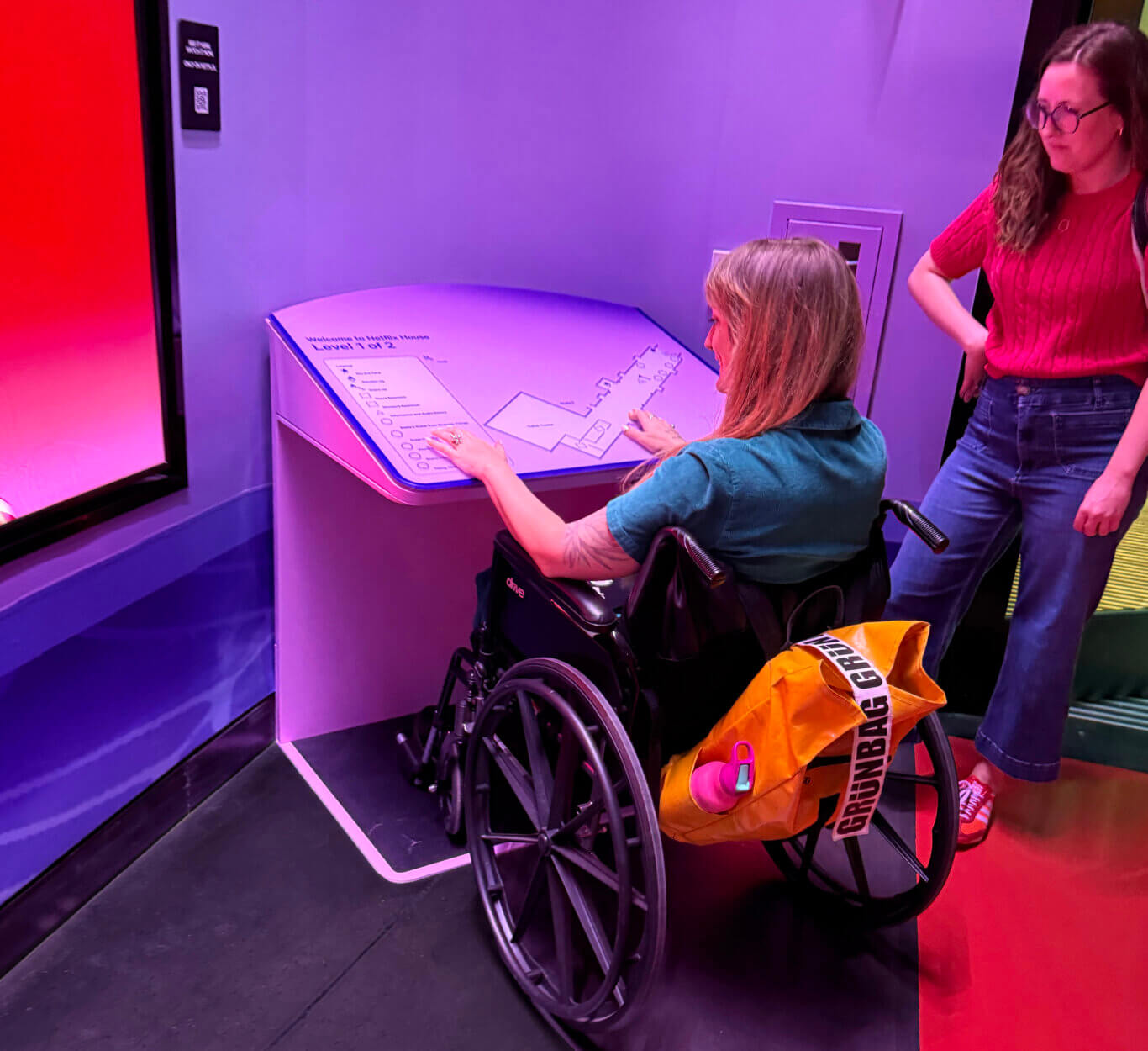Two people interact with an angled tactile floor map of Netflix House Level 1 of 2 set into the corner of a purple lit space. On our left, Robin Marquis, a white nonbinary person in a wheelchair smiles as they touch the legend of the map and the outline of the floor plan. On their left, Maria Braswell, a white woman, stands looking at the map. The map is labeled, "Welcome to Netflix House Level 1 of 2," which shows a floor plan on the right and a legend on the left. A bright red illuminated wall or screen is visible on the left, and a small sign with a QR code is mounted on the wall. An yellow bag with bold lettering hangs on the back of the wheelchair.