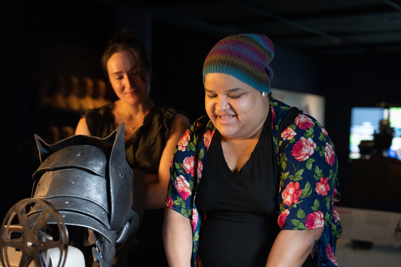 Kate Kita, a white woman with light skin tone, and Nefertiti Matos Olivares, a Latina woman with medium skin tone, interact with a touch table. In the foreground on our left is the back of a dark metallic helmet or armor piece with rivets, and pointed flanges or ears. On our right, Nefertiti touches something out of frame, a large smile on her face, her eyes closed, and a small earbud peeking out of a multicolored striped knit beanie. Behind them, Katie is softly out of focus, looking down toward the object with a slight smile on her face. The background is mostly dark with blurred screens or light sources.