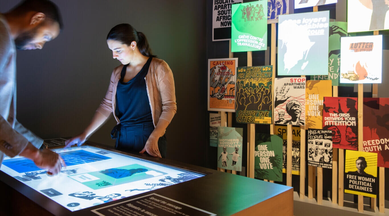 A dimly lit museum or gallery installation where two people are examining a digital interactive display table. One person, with medium skin tone, on the left is blurred in motion while touching the screen, and another with light skin tone stands across the table, focused on the display. To the right, a wall-like arrangement of posters features bold activist graphics and slogans related to democracy, apartheid, labor rights, and political movements.