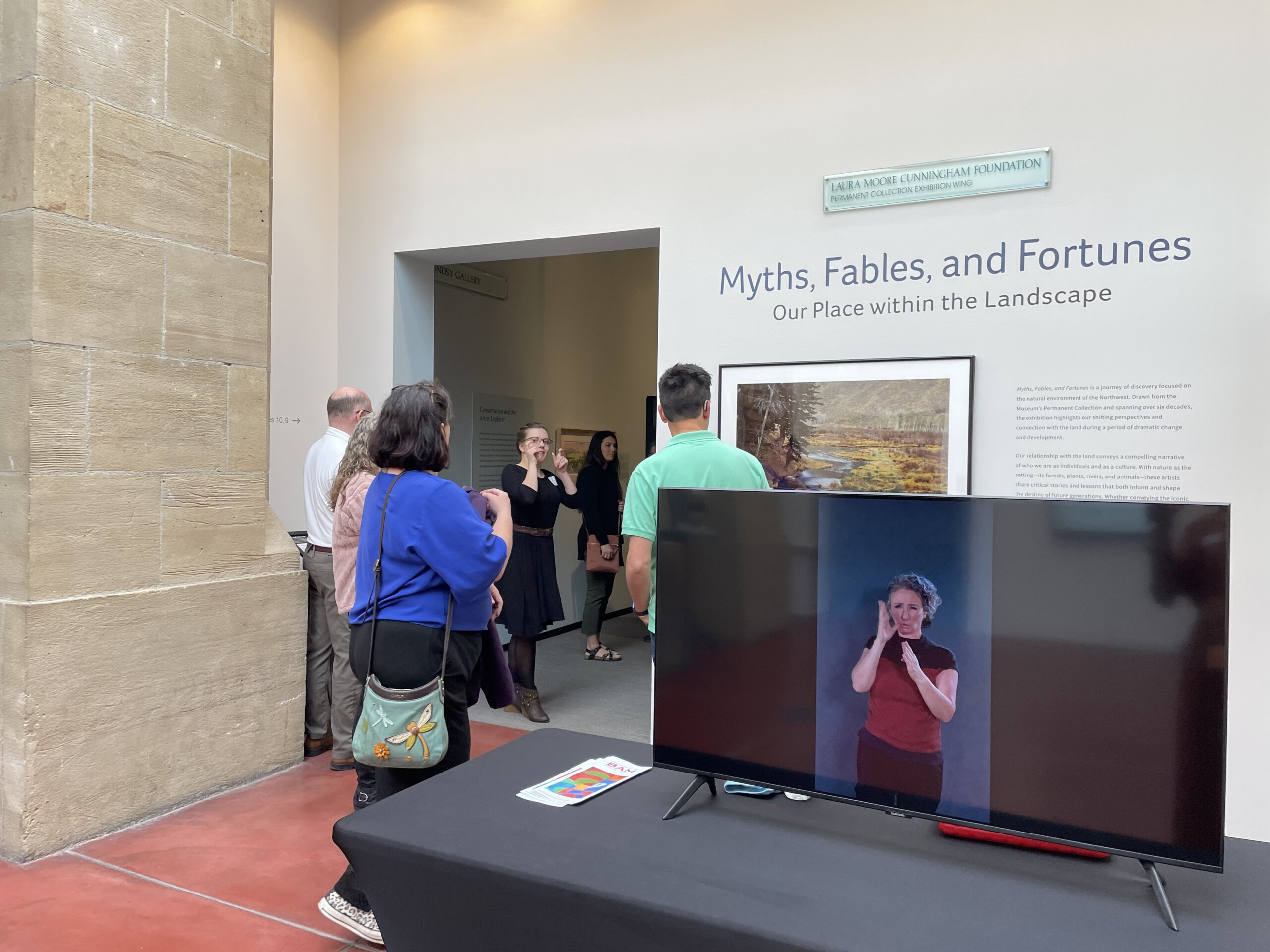 Photograph of a museum lobby during an event. In the foreground, a large flat-screen monitor sits on a black-covered table displaying a white woman signing in ASL against a dark background. Beyond the monitor, several visitors stand in front of a gallery entrance labeled “Myths, Fables, and Fortunes: Our Place within the Landscape,” while a woman in black signs to a small group gathered near the doorway. The space has tall pale stone walls, white gallery surfaces, and reddish polished concrete floors.