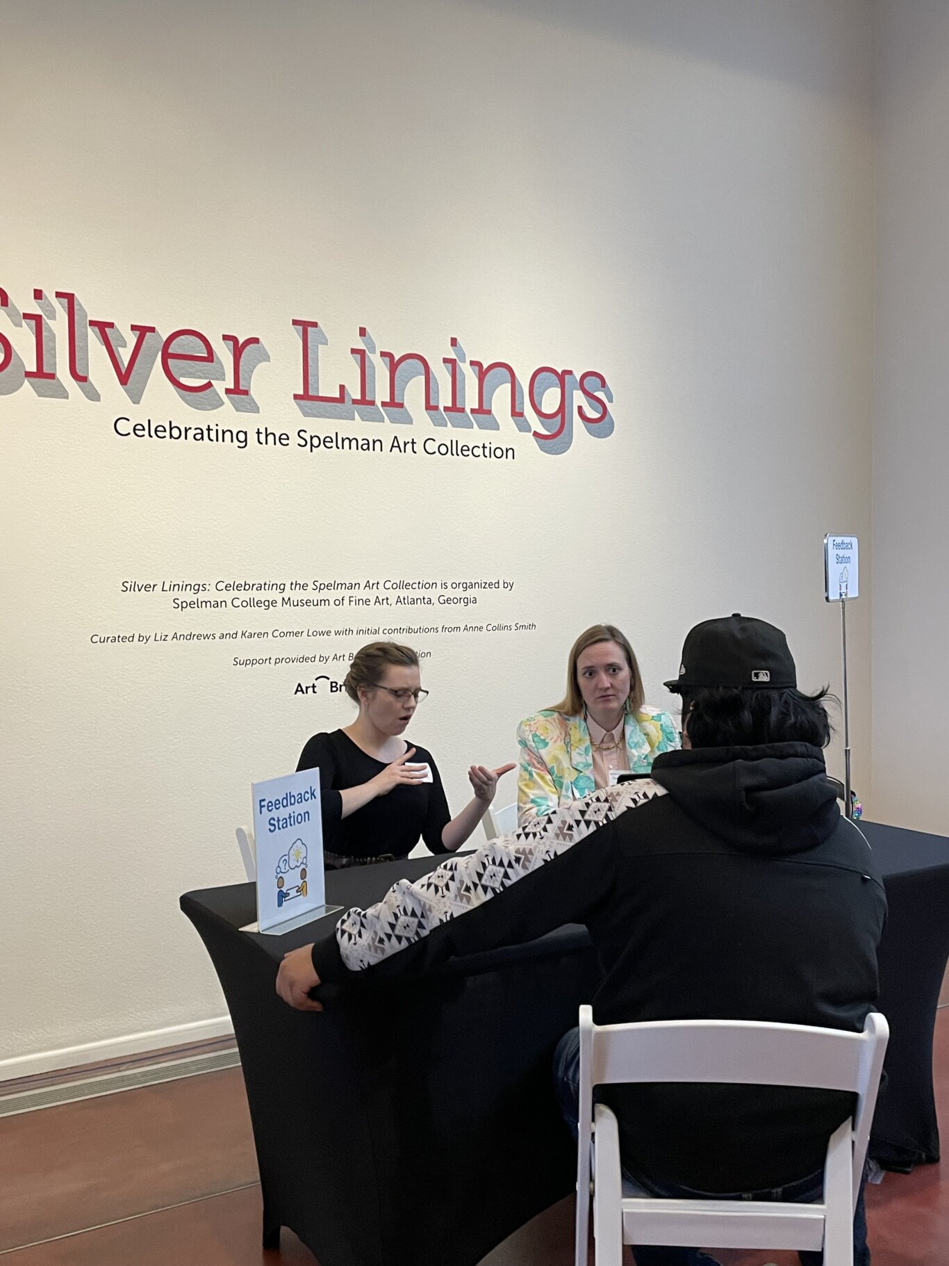 Photograph of a feedback station set up inside a museum gallery. Two people, an white ASL interpreter on our left, and Robin Marquis, a white non binary femme on our left, sit behind a black-covered table signing with a teenage visitor seated across from them with their back to us. The woman on our left signs with her hands while the other looks at the teenager attentively. Small upright signs on the table and behind it read “Feedback Station.” Large wall text behind them reads “Silver Linings: Celebrating the Spelman Art Collection” in layered red and blue lettering.