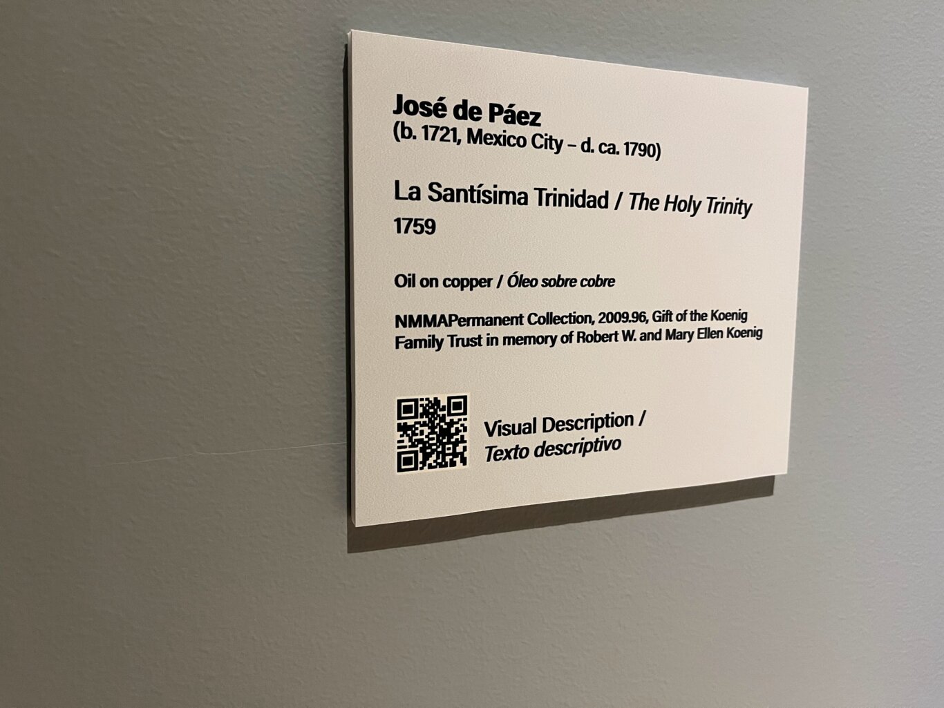 A close-up of a white rectangular museum label attached to a textured gray wall. The Spanish and English bilingual label identifies the artist as "José de Páez (b. 1721, Mexico City - d. ca. 1790)" and the artwork as "La Santísima Trinidad / The Holy Trinity," dated 1759. Near the bottom is a QR code next to the words "Visual Description / Texto descriptivo." It lists the medium as "Oil on copper" and notes it is part of the NMMA Permanent Collection, with a gift credit from the Koenig Family Trust in memory of Robert W. and Mary Ellen Koenig.