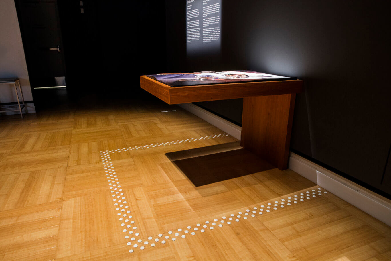 An exhibition space with a table top featuring a barely visible tactile image and white tactile floor markers on a blonde wooden floor. On our right, a wooden display table stickes out from the dark grey wall holding a brightly lit tactile photographic, while a wall panel above it and to the left contains illegible text. The floor is made of warm, light-colored wood parquet and includes rows of raised white tactile markers around the edge of the table.