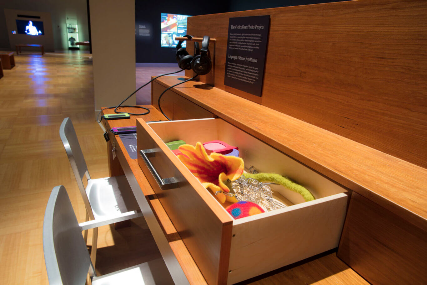 Two metal chairs pulled up to a tactile interactive display table in a warmly lit exhibition space. The long table has a second ledge behind it where a pair of black headphones hangs from a small wooden rod connected by cords to two mobile devices on the tabletop. A sign on the wooden wall reads "The VoiceOverPhoto Project." An open drawer in the foreground contains bright, textured objects, including a felt flower, a wire sculpture, and colorful soft forms. In the background, the gallery continues with polished wood floors, benches, wall displays, and a glowing video screen.