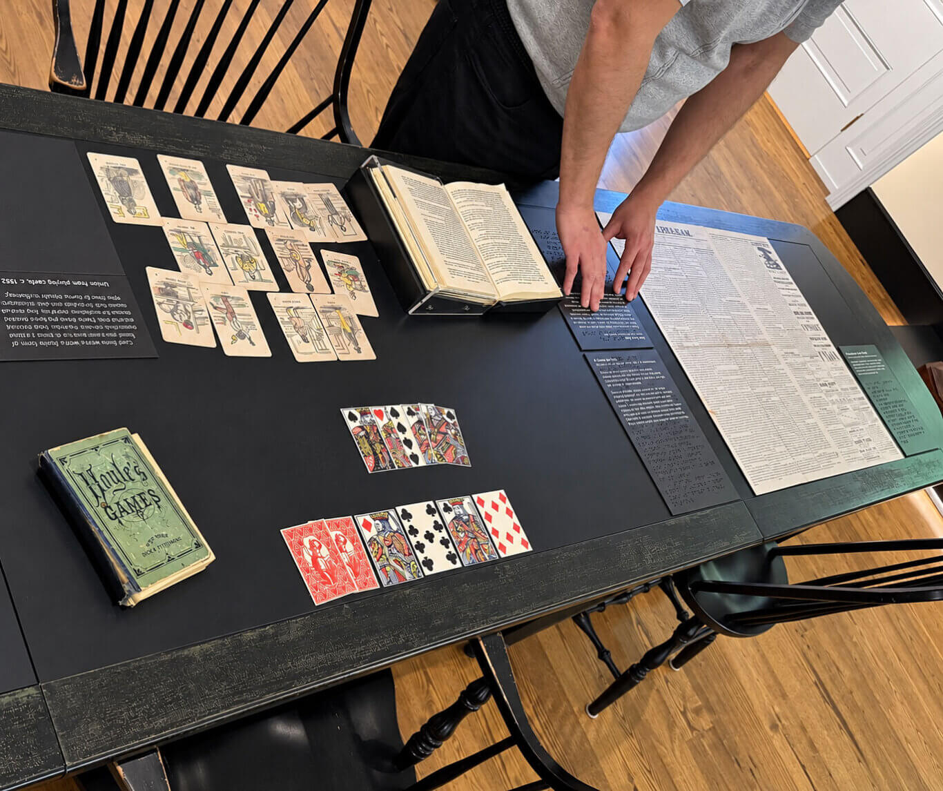 Viewed from overhead and at an angle, a person with light skin tone leans over a large black wooden table, touching an exhibit of historical card games. Their head is cropped out, and both hands rest near a group of dark interpretive panels with printed white text and Braille. At our upper left, about fifteen aged illustrated cards are arranged in loose rows beside an upside-down black label whose heading reads "Union Playing Card Co. 1882." Near the top center, a yellowed book lies open on a black display cradle. Several traditional playing cards are spread faceup across the center and lower center, including black spades and clubs, red diamonds and hearts, court cards, and two red-backed cards. At lower left, a worn green book cover reads, from top to bottom, "Hoyle's," "GAMES," "EDITED BY," and "DICK & FITZGERALD." On the right, a large cream-colored broadsheet contains tightly packed columns and advertisements; its masthead is partly covered and blurred. Additional blue-gray and green interpretive panels surround it, combining small printed text with lines of raised Braille. Black spindle-back chairs surround the table, and a honey-colored wooden floor and white paneled cabinets fill the background.