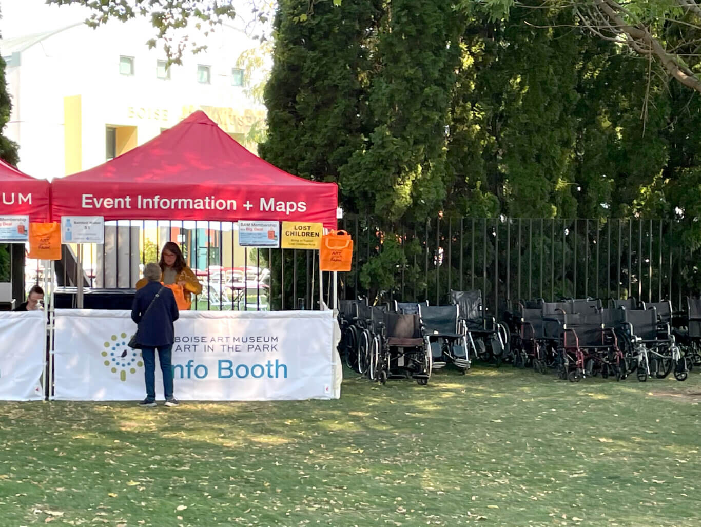 An outdoor event information booth set up on a grassy lawn. A bright red canopy reads "Event Information + Maps," and a banner on the front says "Boise Art Museum Art in the Park Info Booth." A few people are standing or sitting at the booth, with orange bags and signs visible, including one for "Lost Children." To our right, many wheelchairs are lined up in rows near a metal fence beneath large leafy trees.