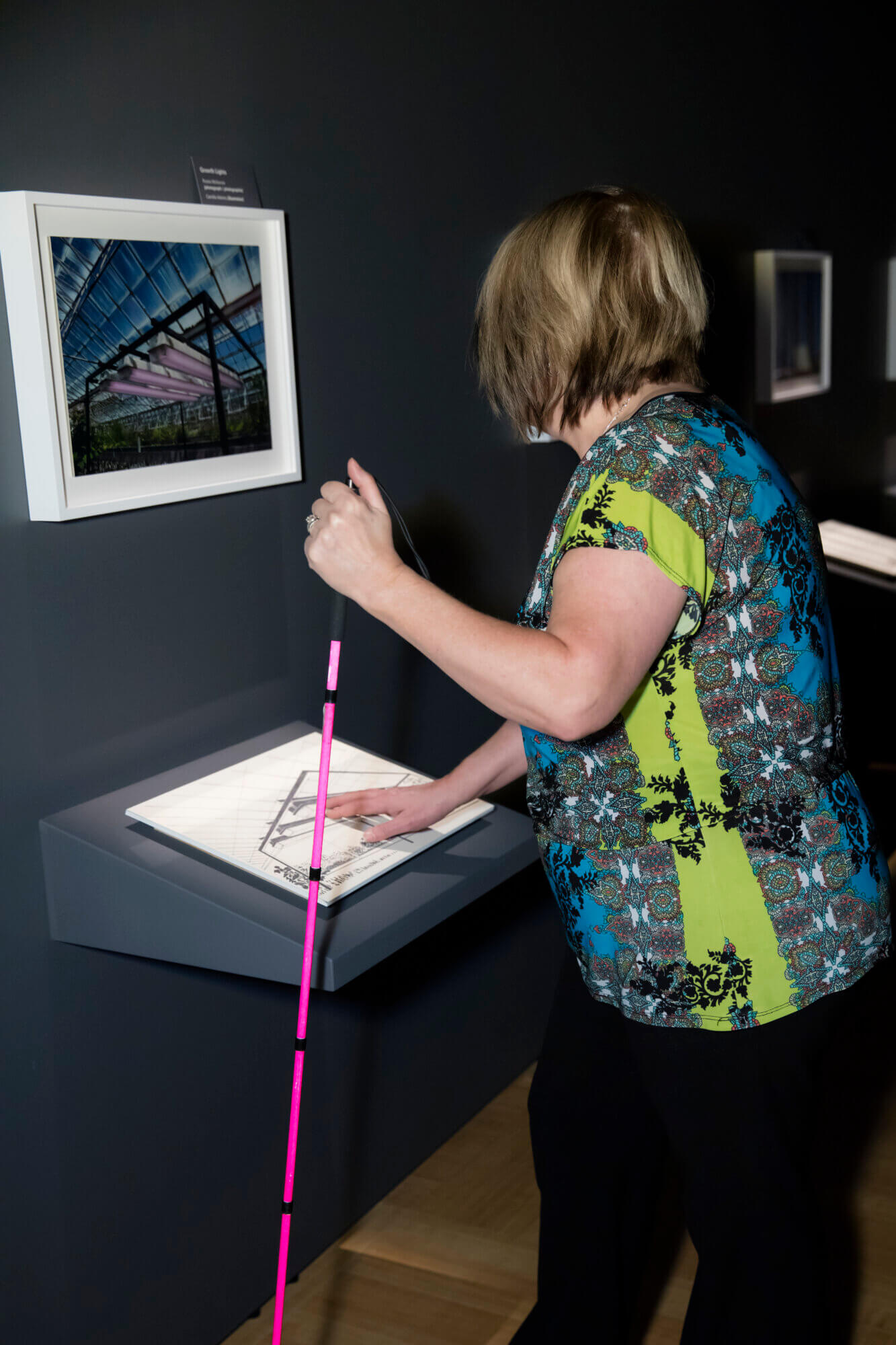 A person with pale skin standing in a dimly lit exhibition space, viewed mostly from behind and the side. She is holding a bright pink long cane in one hand and placing her right hand on a raised tactile image of a simplified black and white glass roofed structure or greenhouse interior displayed on a tilted ledge. In front of her, on the dark grey wall, is a color photograph, framed in white, of the same scene. Additional framed works are visible blurred in the background.