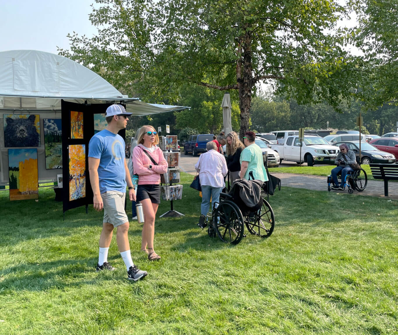 A sunny outdoor art market set up in a park. On our left, a white tent displays colorful paintings, including a large bear portrait and landscape-style artwork. Several people with light skin tone are walking on the grass in front of the booth. An additional cluster of people look at the art in the booth next to a parked wheelchair. In the background, there are more visitors, parked cars, a closed patio umbrella, and large leafy trees providing shade.