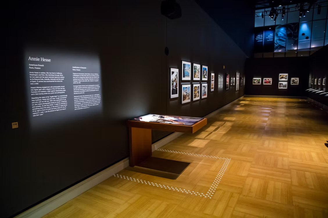 A dimly lit photography gallery displays framed color photographs along dark walls, with a wooden interactive table projecting from the left wall and white tactile dots outlining a path across the parquet floor. A large illuminated bilingual wall label on the left reads "Annie Hesse," followed by "American/French" and "Paris, France" in English and French followed by illegible wall text. A small yellow marker below the label reads "116." Farther along the left wall, photographs of people and outdoor scenes hang in white mats and dark frames. More framed photographs line the distant back and right walls. High above them, blue illuminated panels include a large close-up photograph of a person's face and several blocks of white text that are too small and blurred to read. Overhead spotlights and the display table cast warm rectangular reflections and long shadows across the honey-colored wooden floor.