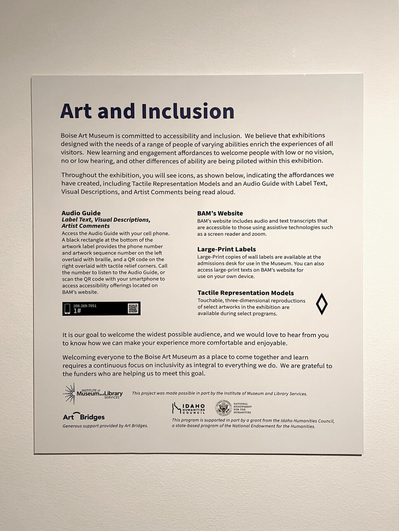 A rectangular white museum wall sign with black text is mounted on a textured off-white wall; it is titled Art and Inclusion and explains accessibility offerings at the Boise Art Museum, with two columns of information featuring an Audio Guide, BAM's Website, Large-Print Labels, and Tactile Representation Models. Underneath that are a small sample audio-guide label, a tactile-representation icon, and funder logos near the bottom. The visible text reads: Art and Inclusion. Boise Art Museum is committed to accessibility and inclusion. We believe that exhibitions designed with the needs of a range of people of varying abilities enrich the experiences of all visitors. New learning and engagement affordances to welcome people with low or no vision, no or low hearing, and other differences of ability are being piloted within this exhibition. Throughout the exhibition, you will see icons, as shown below, indicating the affordances we have created, including Tactile Representation Models and an Audio Guide with Label Text, Visual Descriptions, and Artist Comments being read aloud. In the left column, the heading reads Audio Guide, followed by italic text: Label Text, Visual Descriptions, Artist Comments. The paragraph below reads: Access the Audio Guide with your cell phone. A black rectangle at the bottom of the artwork label provides the phone number and artwork sequence number on the left overlaid with braille, and a QR code on the right overlaid with tactile relief corners. Call the number to listen to the Audio Guide, or scan the QR code with your smartphone to access accessibility offerings located on BAM's website. A black sample label below includes a phone icon, the number 208-269-7051, the sequence 1, and a QR code. In the right column, the heading reads BAM's Website, followed by: BAM's website includes audio and text transcripts that are accessible to those using assistive technologies such as a screen reader and zoom. Below that, another heading reads Large-Print Labels, followed by: Large-Print copies of wall labels are available at the admissions desk for use in the Museum. You can also access large-print texts on BAM's website for use on your own device. A third heading reads Tactile Representation Models, followed by: Touchable, three-dimensional reproductions of select artworks in the exhibition are available during select programs. A black outlined diamond-shaped icon appears beside this section. Across the lower middle, larger text reads: It is our goal to welcome the widest possible audience, and we would love to hear from you to know how we can make your experience more comfortable and enjoyable. Below it, text reads: Welcoming everyone to the Boise Art Museum as a place to come together and learn requires a continuous focus on inclusivity as integral to everything we do. We are grateful to the funders who are helping us to meet this goal. At the bottom left, a logo reads INSTITUTE of Museum and Library SERVICES, with adjacent text: This project was made possible in part by the Institute of Museum and Library Services. Below left, another logo reads Art Bridges, followed by: Generous support provided by Art Bridges. Near the lower center, a logo reads IDAHO HUMANITIES COUNCIL, next to a seal and text reading NATIONAL ENDOWMENT FOR THE HUMANITIES. The final line reads: This program is supported in part by a grant from the Idaho Humanities Council, a state-based program of the National Endowment for the Humanities.
