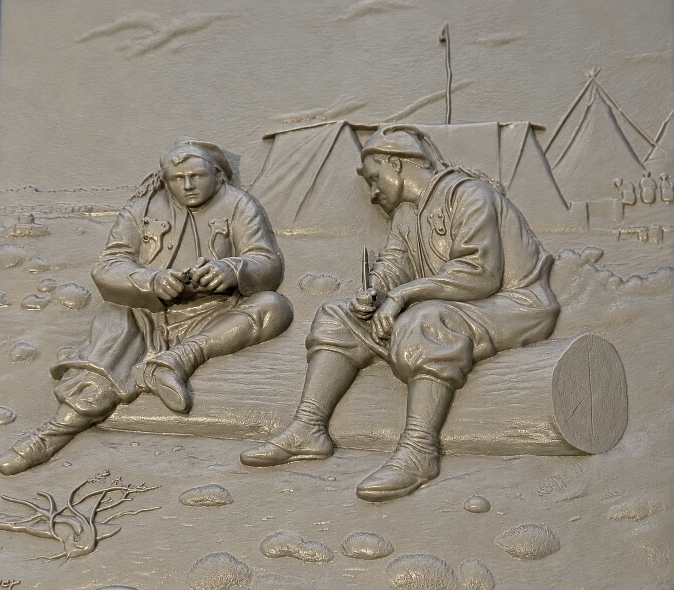 A monochrome gray tactile relief depicts two soldiers resting in a sparse encampment, whittling wood. On our left, the man sits on the ground with one leg extended and the other bent across the body. He wears a soft cap or head covering, a heavy decorated coat, loose trousers, and ankle-high footwear, and he holds a small indistinct object between both hands while looking downward. On our right, the man sits hunched forward on a horizontal log, wearing a headband or close-fitting cap, a similarly decorated coat, loose knee-length trousers, wrapped lower legs, and soft shoes. He holds a narrow pointed tool upright in one hand and steady another small object near their lap. The circular cut end of the log faces us at the far right and is incised with tree-ring and crack patterns. Several angular canvas tents rise across the background, with poles and taut seams rendered in shallow relief. A partial raised marking on the tent at the right edge is cropped. Scattered stones, tufts of grass, and a bare branching limb cover the foreground, while faint birds or clouds appear in the shallowly textured sky.