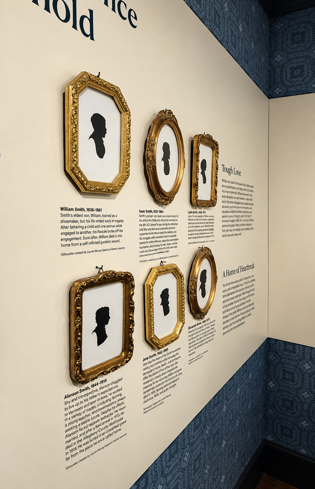 A museum exhibition displays six black profile silhouettes in ornate gold frames on a cream wall, arranged in two rows of three with the headings "Tough Love," and "A Home of Heartbreak." The frames vary in shape, including octagonal, oval, and rectangular designs, and each portrait has a biographical label beneath or beside it. Dark blue wallpaper with repeating floral and geometric patterns covers the sloping ceiling and the lower-right wall. The names of the two portraits on our left are visible, "William Smith, 1836-1861, " and "Alanson Smith, 1844-1914."