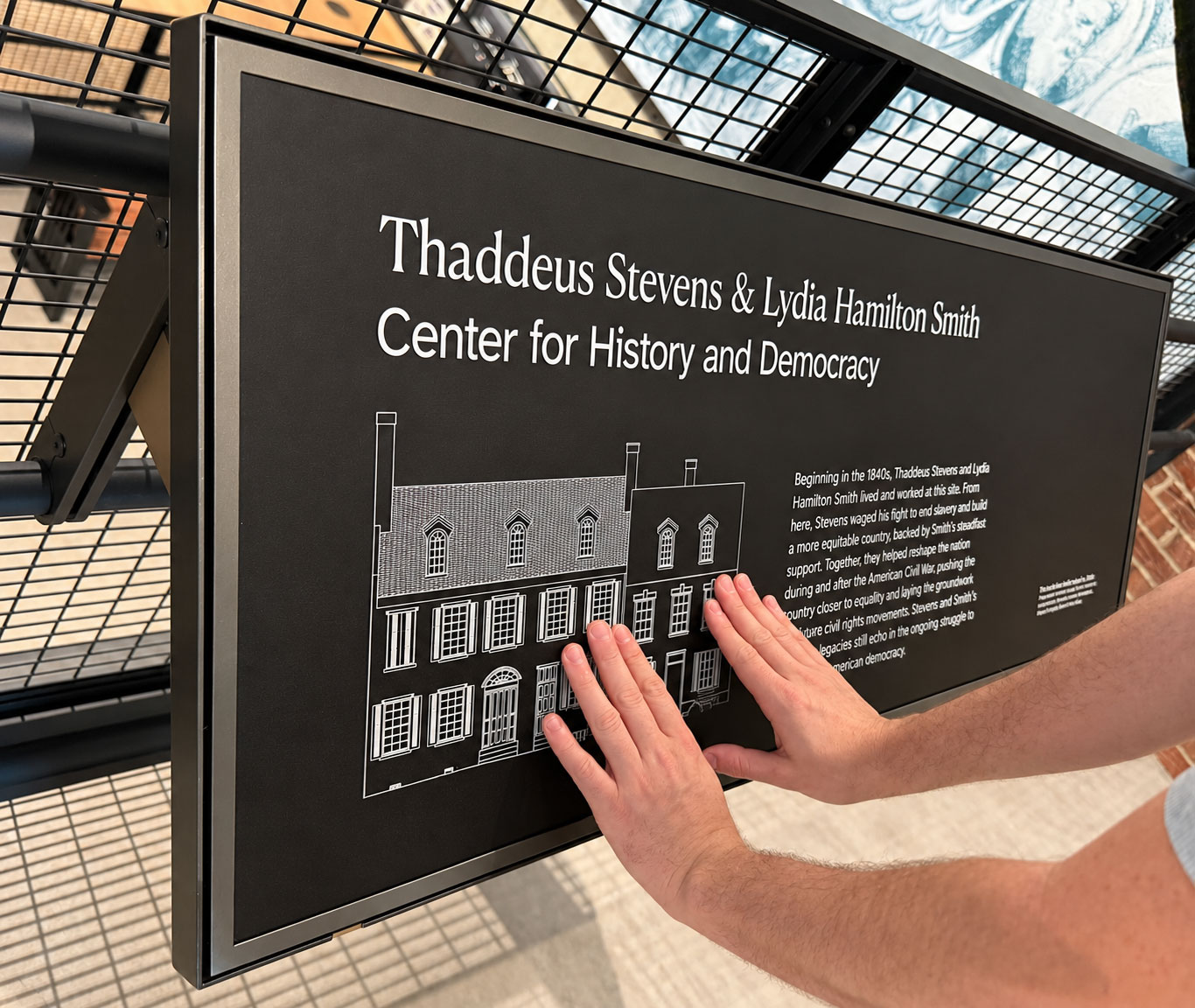A black tactile interpretive panel mounted on an angled metal grid bears raised white lettering and a line drawing of a historic building, the "Thaddeus Stevens & Lydia Hamilton Smith Center for History and Democracy." Two light-skinned hands reach in from our lower right and touch the building illustration. Underneath the title, is the line drawing of the house and text about the house. The white architectural drawing shows a connected row of multi-story brick buildings with chimneys, dormer windows, shutters, doors, and many rectangular windows.