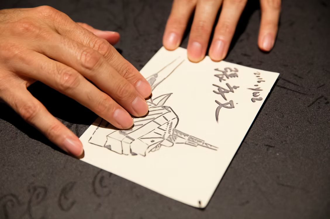 A close-up color photograph shows two light toned hands holding a cream-colored postcard or drawing card diagonally against a dark, subtly patterned tabletop. The left hand spreads across the lower-left portion of the card, partially covering a fine black-and-gray drawing of an angular architectural shape with long pointed forms extending above and to the right. The other hand steadies the card along its upper edge. Large, dark handwritten Japanese characters appear near the upper-right area.