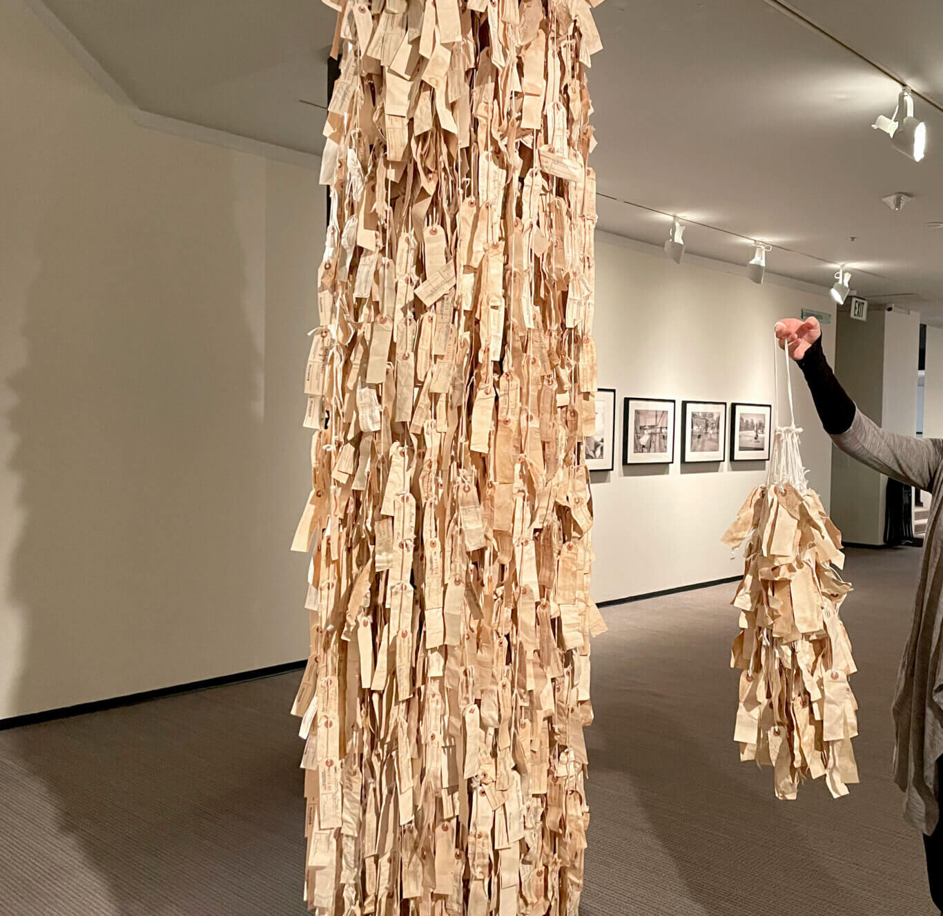 A gallery installation fills the center of the image: a tall hanging column densely covered with cream paper tags dangling from white strings. The tags overlap in thick vertical layers, many with reinforced circular holes in reddish brown and loops of cord tied through them, creating a shaggy, curtain-like surface. Some tags contain printed boxes, handwriting, or stamped marks, but the text on them is too small and blurred to read. On our right, a cropped with light skin tone wearing a gray short-sleeved top over a black long-sleeved layer holds up a white cord attached to a smaller matching bundle of dangling tags. The room has gray carpet, pale walls, and a ceiling with bright track lights aimed at the artworks. In the background on the right wall, several framed black-and-white images hang in a row.